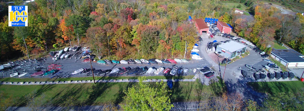 Aerial view of Off Shore Marine dealership showing main building, parking lot with several boats on display, surrounded by green trees and open sky, conveying a welcoming and professional atmosphere